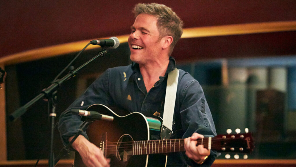 Josh Ritter performs live. Rooted & Restless Issue 3 features musician playing guitar and singing into a microphone.