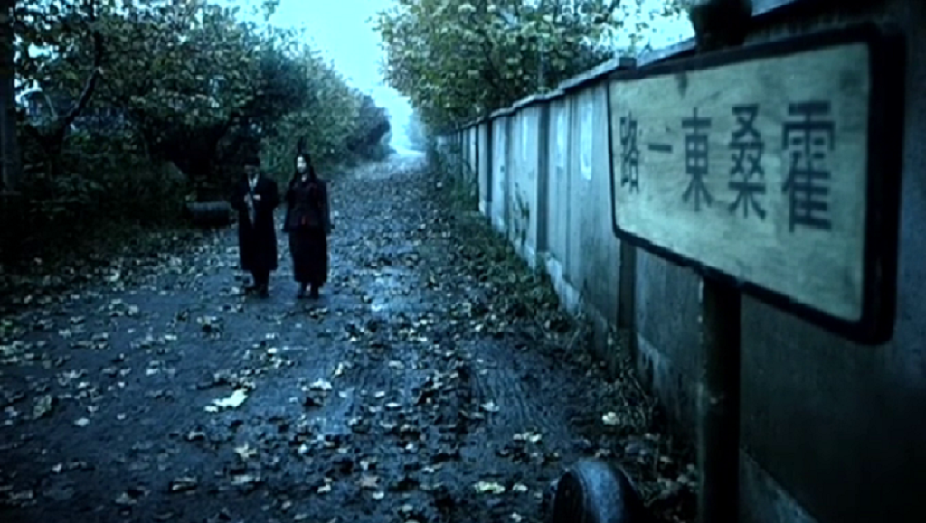Lou Ye film still. Two figures walk along a path next to a wall with a sign in Chinese characters. Don't Be Young.