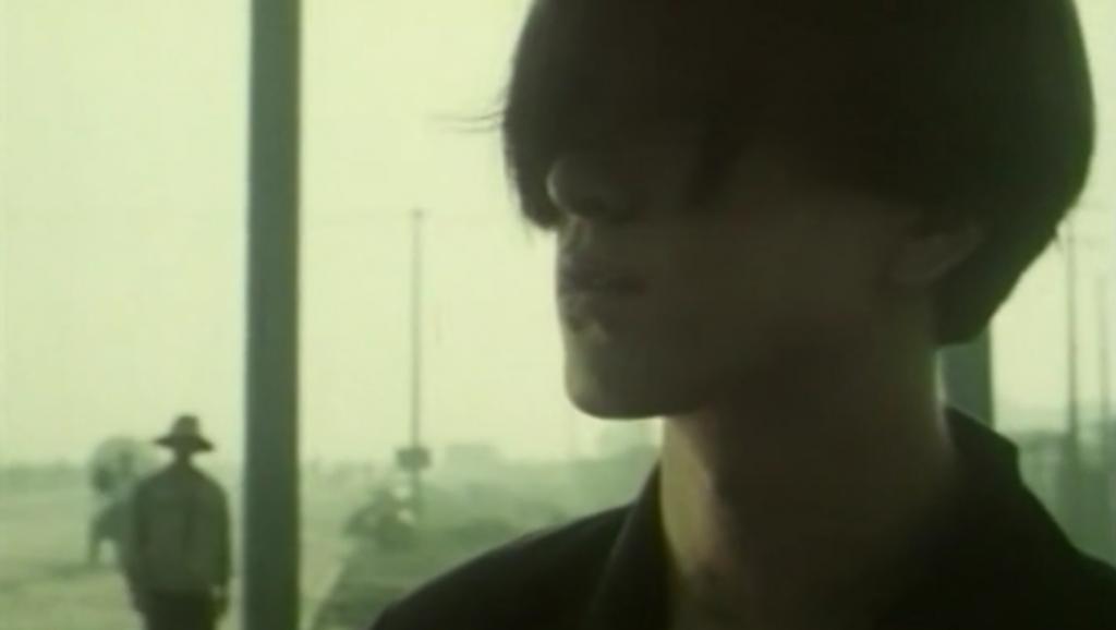 Lou Ye's Weekend Lover movie still. Close-up of a person with dark hair. A figure in a hat stands in the background.