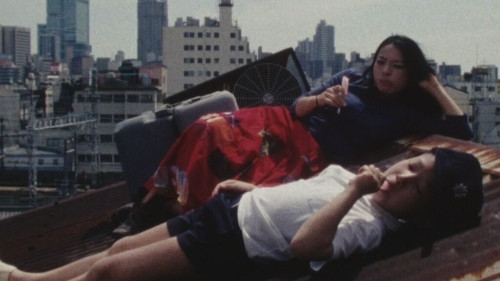 Film still from The Kamagasaki Cauldron War featuring two people relaxing on a rooftop with a cityscape backdrop.