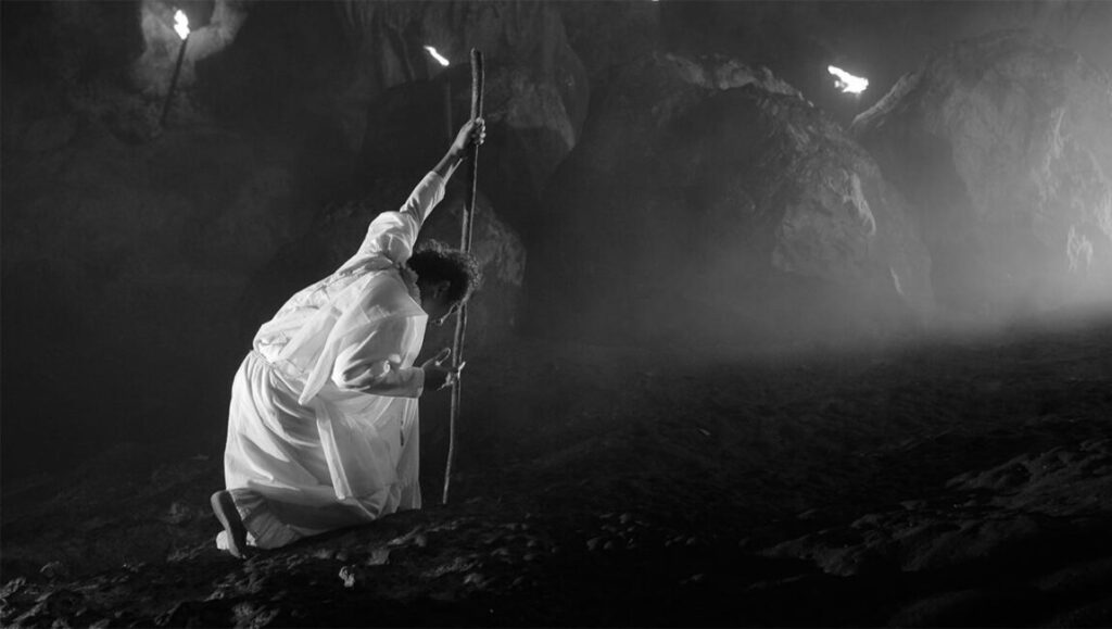 Black and white still from Lav Diaz's A Lullaby to the Sorrowful Mystery featuring a kneeling figure with a staff.