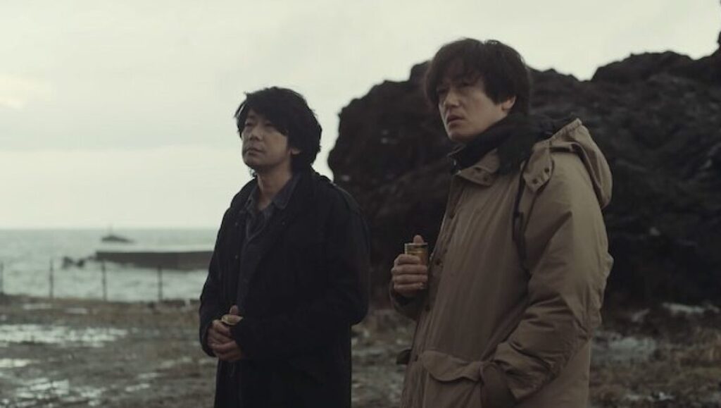 Red Snow film still: Two men stand on a rocky shore, gazing out at the sea. Japanese cinema.