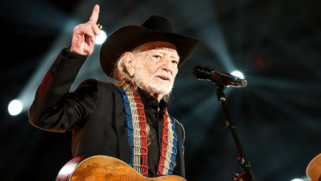Willie Nelson performing live. Country music icon with guitar, hat, and signature braids. Rooted & Restless.