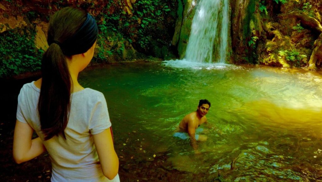Yara film still: Woman watches man swimming in a pool below a waterfall, surrounded by lush greenery.