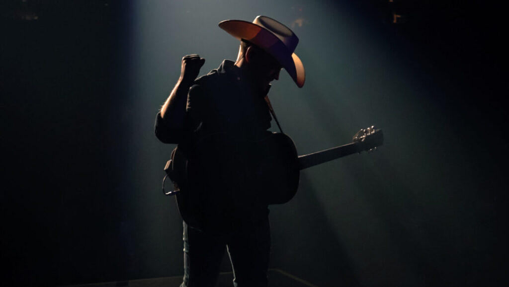 Rooted & Restless: Silhouette of musician with guitar and cowboy hat in spotlight. Issue 5.