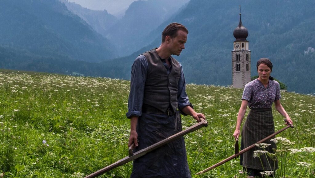 A Hidden Life movie scene. A man and woman mow a field in front of mountains and a church tower. Toronto International Film Festival.
