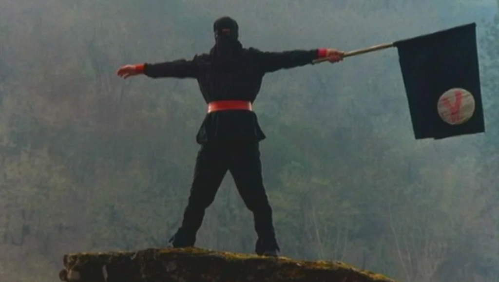 Summer Blockbuster!?! | Episode 105: Gymkata Gymkata movie scene. Ninja on a rock holding a flag. Summer Blockbuster episode. Martial arts film.