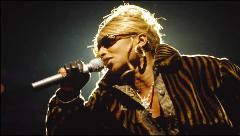 Mary J. Blige performing My Life. Singer in sunglasses, patterned coat, and jewelry holding a microphone.