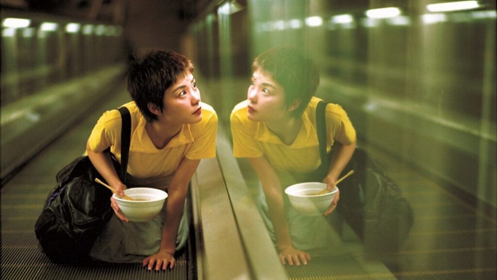 Chungking Express film still. Woman with bowl and chopsticks on an escalator, reflected in glass. March 2020 film review.