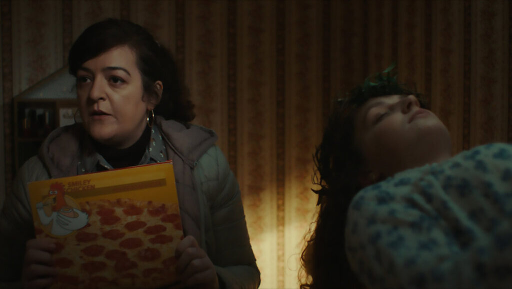 Extra Ordinary movie scene: Woman holds pizza box, another lies down. Comedy film by Mike Ahern & Enda Loughman.