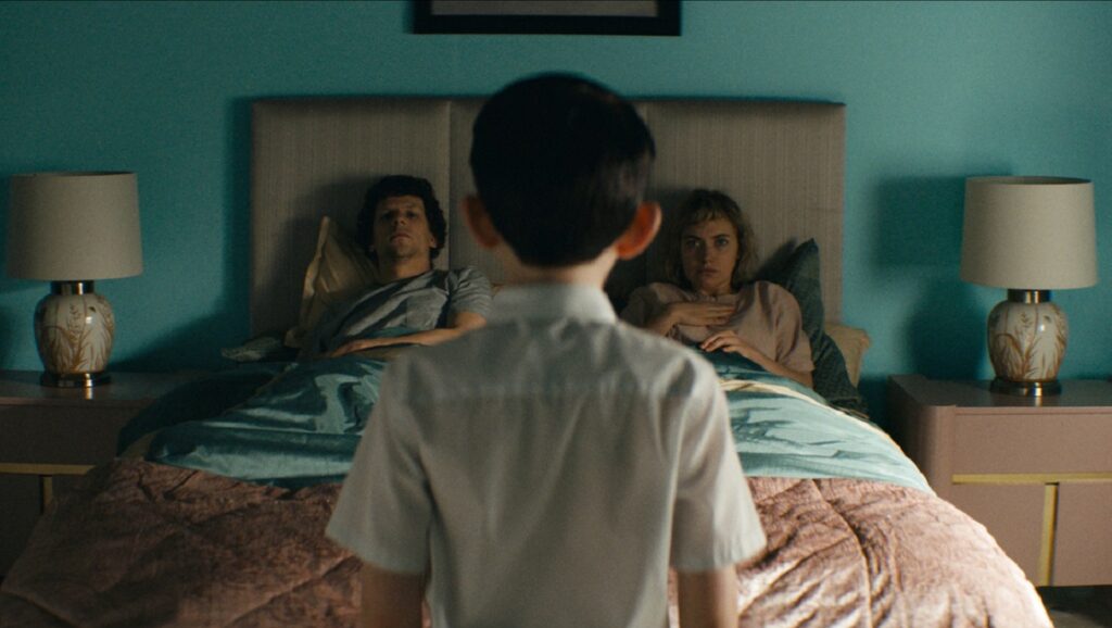 Vivarium movie scene. A couple lies in bed as a child stands at the foot. Starring Imogen Poots and Jesse Eisenberg.