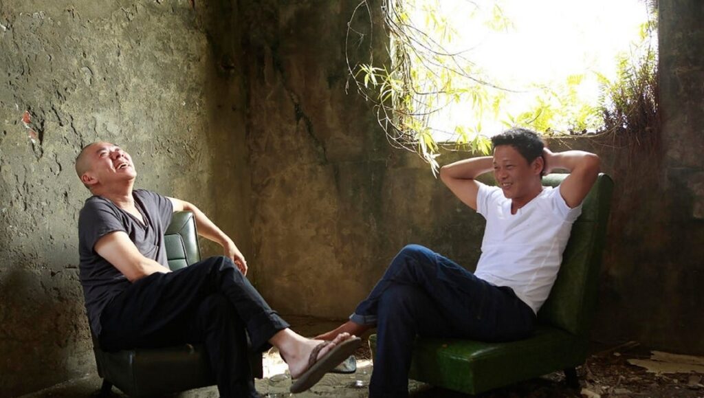 Tsai Ming-liang's Afternoon film still. Two men sit in chairs, one laughing. Ruined building backdrop. Afternoon mood.