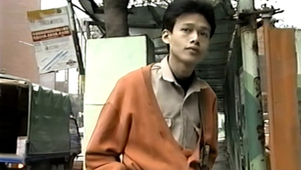 A scene from Boys by Tsai Ming-liang. Young man in orange cardigan stands in an alleyway.