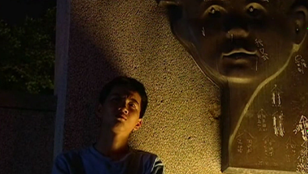 My Stinking Kid | Tsai Ming-liang My Stinking Kid film still: Man leaning against a wall with a large, engraved face. Tsai Ming-liang film.