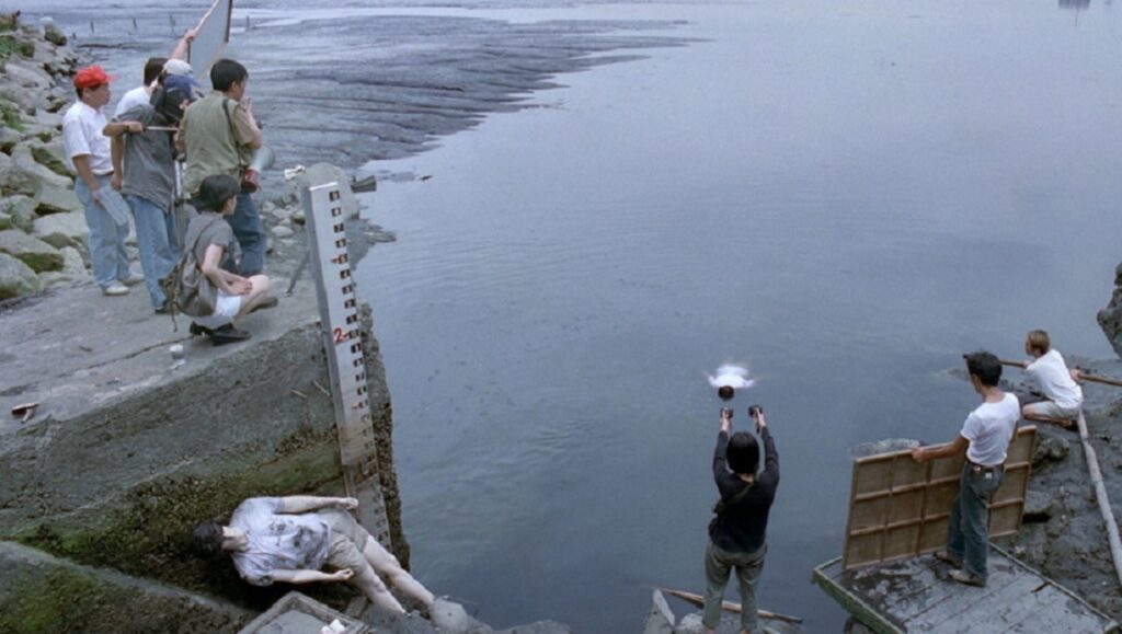 Tsai Ming-liang's The River scene. People near water, one throws a ball. Film still.