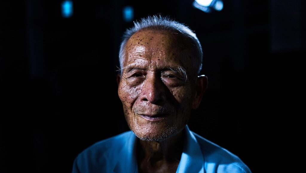 Close-up of elderly man's face, Tsai Ming-liang's Your Face film. Wrinkled skin, gray hair, and dark background.