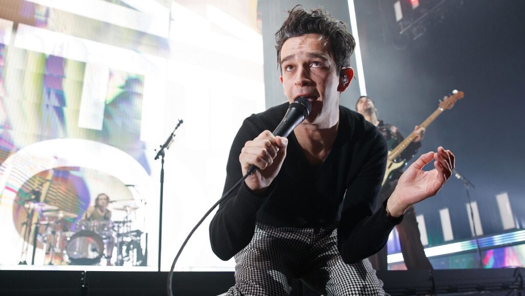 The 1975 | Notes on a Conditional Form The 1975 performing live. Singer Matty Healy sings into a microphone on stage with his band. Pop music concert.