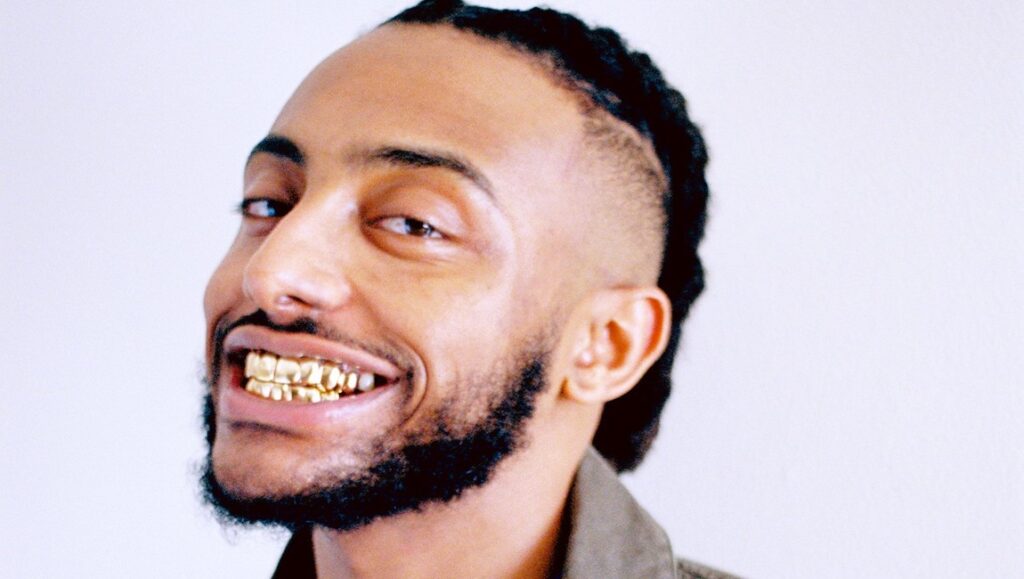 Aminé | OnePointFive Aminé, the artist behind *OnePointFive*, smiling with gold teeth. Braided hair and a trimmed beard.
