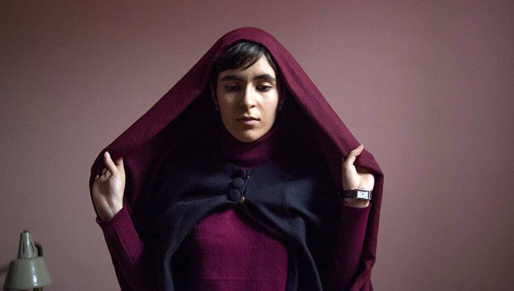 Ava film still. Sadaf Foroughi directs a young woman in a maroon hooded cloak and turtleneck sweater. Portrait.
