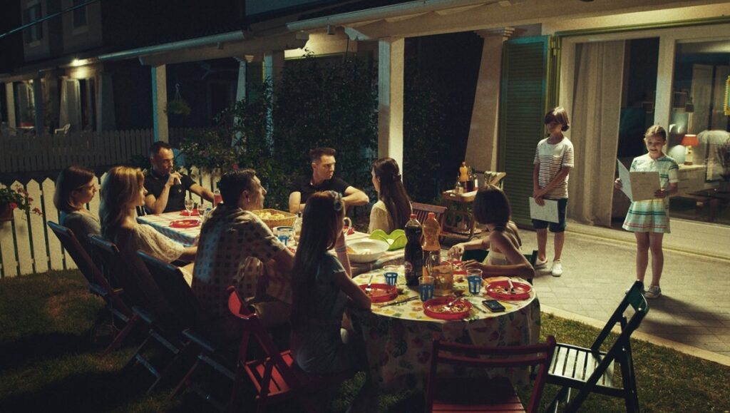 Bad Tales | Damiano and Fabio D’Innocenzo Bad Tales film scene: Family dinner party at night. Children stand with papers.