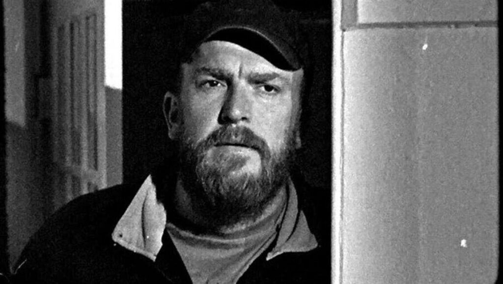 Black and white still from Bait by Mark Jenkin. Bearded man in a cap looks out from a doorway. Indie film.