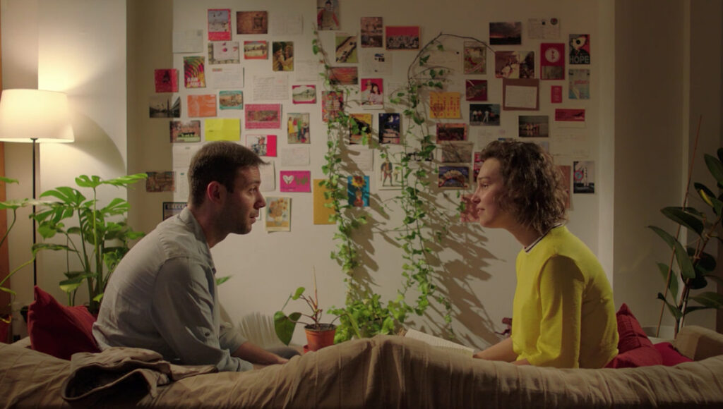 Belonging | Burak Çevik Belonging film scene. Couple sitting face to face in a room with plants and postcards on the wall.