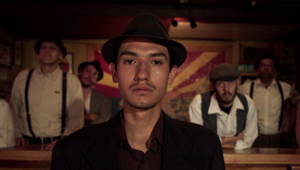 Bisbee '17 film still. Men in vintage clothing and hats stand in a dimly lit room with an Arizona flag.