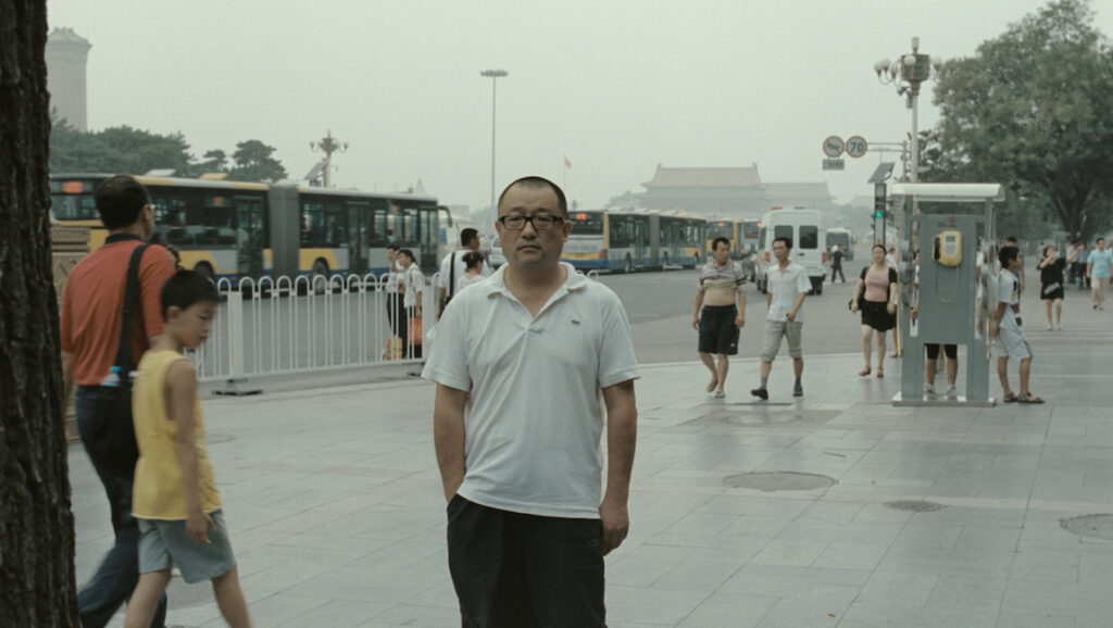 Chinese Portrait | Wang Xiaoshuai Chinese Portrait. Wang Xiaoshuai film still of a man standing in a busy urban Beijing street with buses and pedestrians.