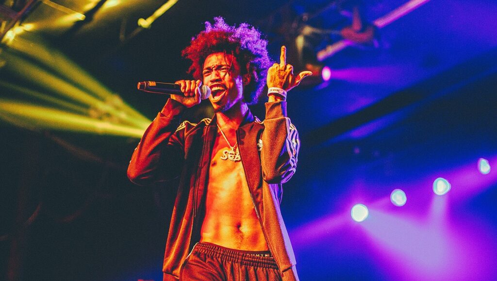 DaBoii | Neva Lookin Back DaBoii performing live. Rapper with afro hairstyle, holding a microphone, flipping the bird on stage. Neva Lookin Back.