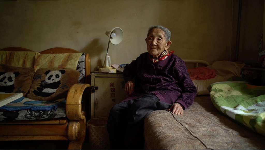 Still from Wang Bing's Dead Souls documentary. Elderly woman sits in a dimly lit room. Panda pillows on a couch.