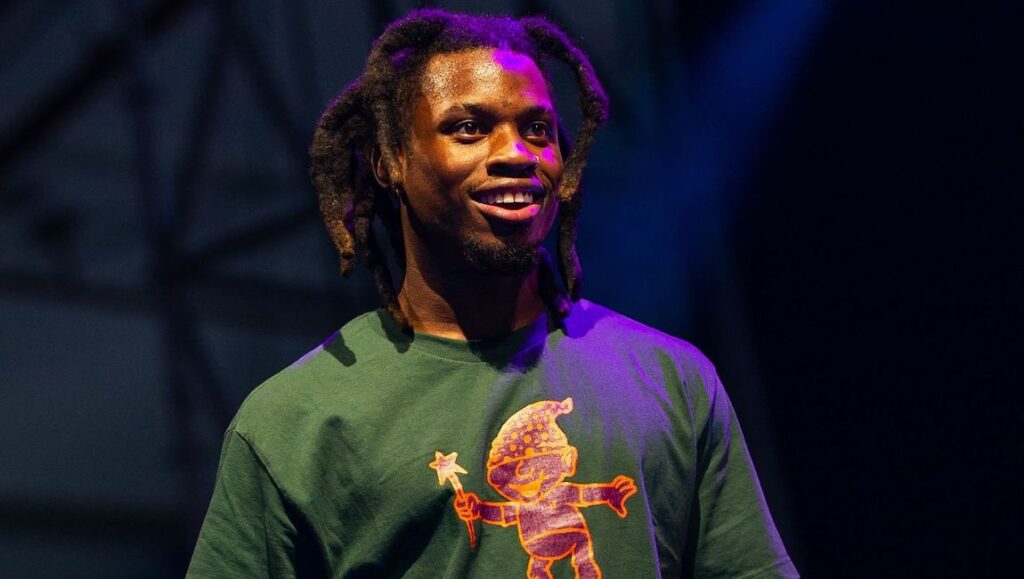 Denzel Curry | TA1300 Denzel Curry, TA1300 era. Rapper with dreadlocks, wearing a green t-shirt with a cartoon graphic, smiling.