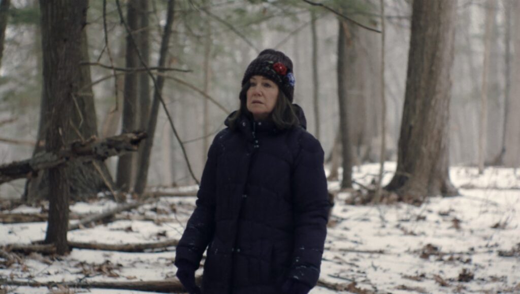 Diane in winter woods. Kent Jones film. Woman in hat and coat.
