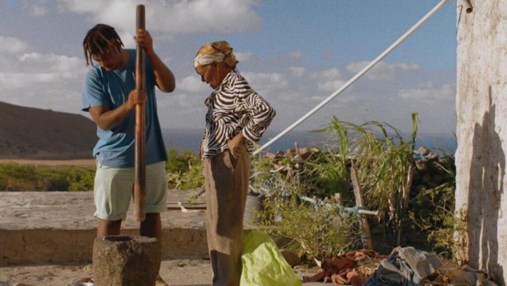 Djon Africa | João Miller Guerra & Filipa Reis Djon Africa film still. Man pounding grain with elder woman in zebra print nearby. Rural, sunny scene.