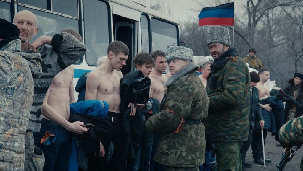 Scene from Donbass film: Men, some shirtless, stand near a bus. A flag of the Donetsk People's Republic waves.