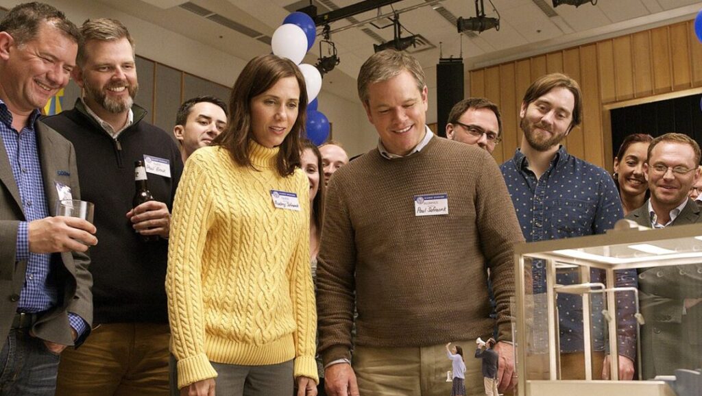 Downsizing film still. Kristen Wiig and Matt Damon look at a model with tiny people. Crowd in the background.