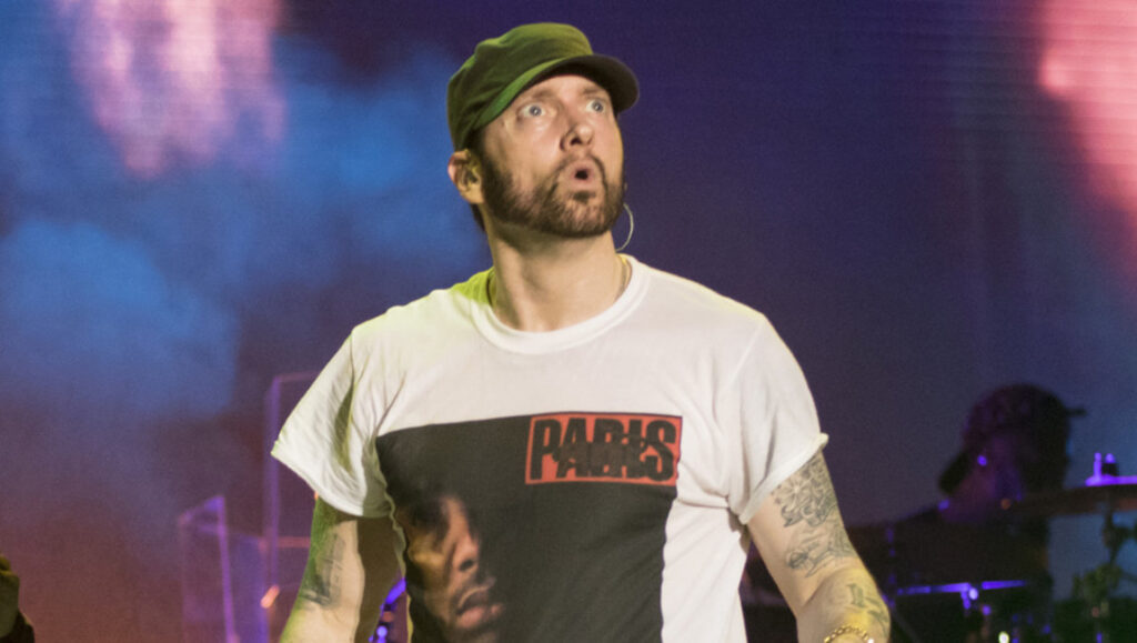 Eminem performing live. Rapper in a hat and Paris t-shirt during the Kamikaze era. Concert stage lighting.