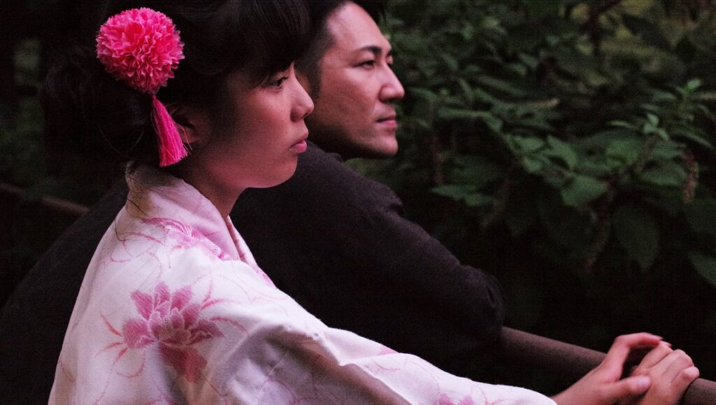 Family Romance, LLC film still. Woman in kimono with pink flower, man beside her. Werner Herzog film.