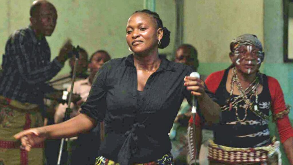 Félicité | Alain Gomis Félicité film still. A woman sings into a microphone, surrounded by musicians and a performer with face paint.