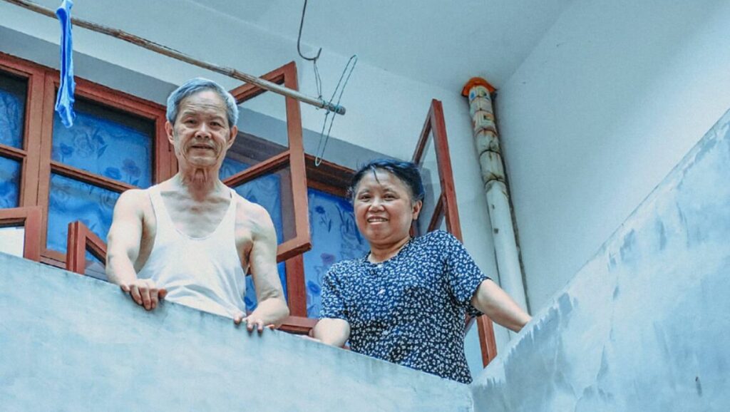 Four Springs | Lu Qingyi Lu Qingyi's Four Springs: Elderly couple smiling on balcony. Man in tank top, woman in floral blouse. Open window.