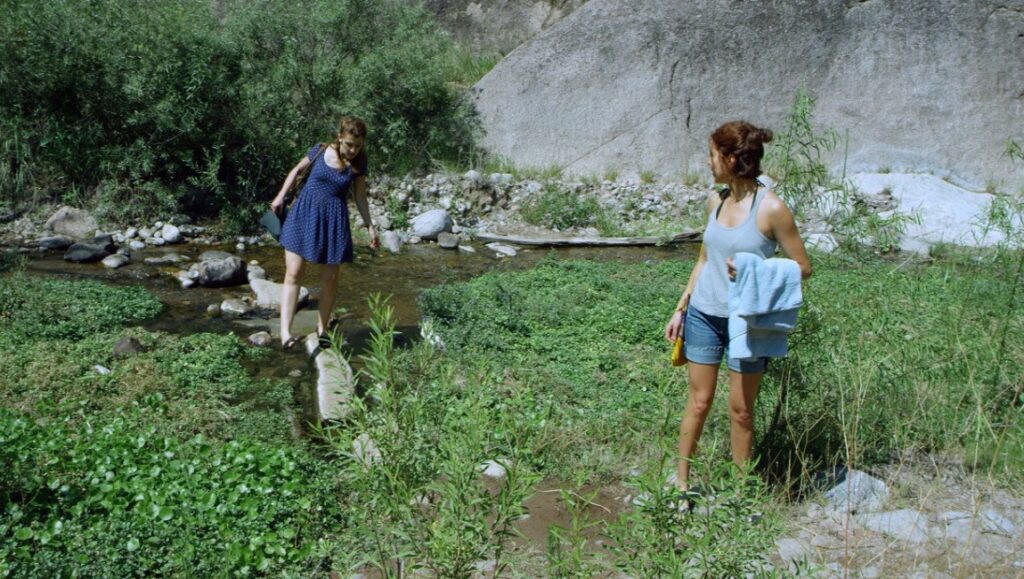 Isabella film scene: Two women near a stream. One crosses the water, the other watches. Summer day in nature.