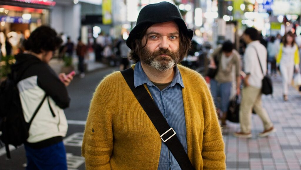 Jim O'Rourke in Tokyo. Musician wearing a black hat and yellow cardigan. Shutting Down Here album era.