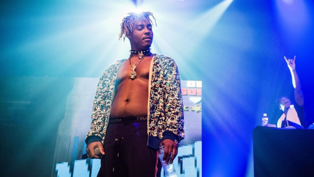Juice WRLD | Goodbye & Good Riddance Juice WRLD performing live. Rapper on stage with dreadlocks, jewelry, and patterned jacket. Concert with blue lighting.
