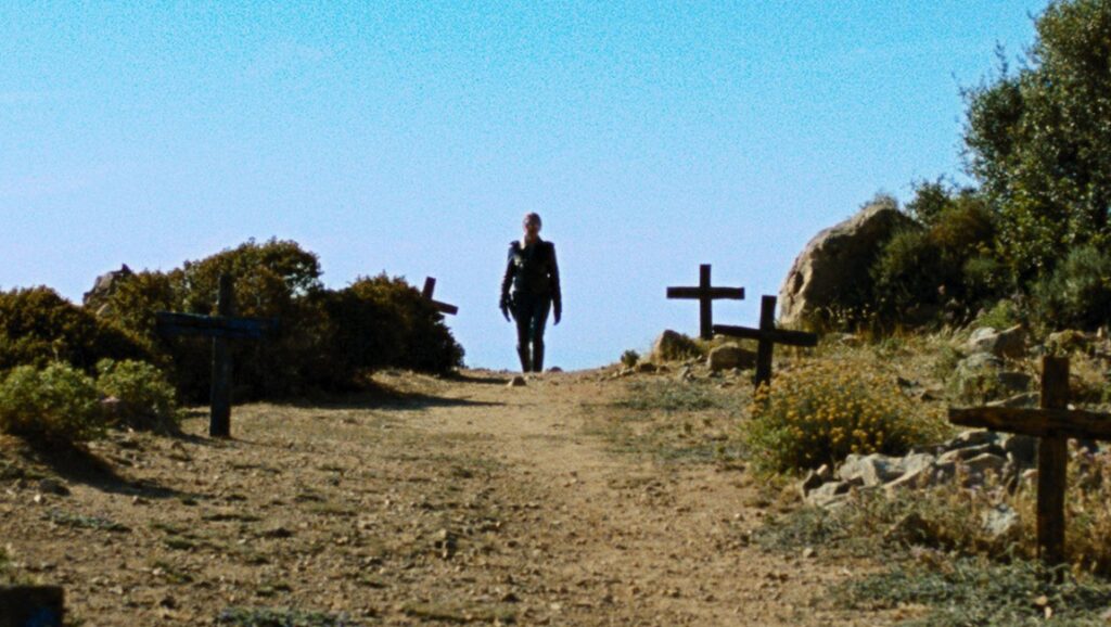 Let the Corpses Tan | Hélène Cattet & Bruno Forzani Let the Corpses Tan film still. Figure walks a path lined with crosses under a clear blue sky.