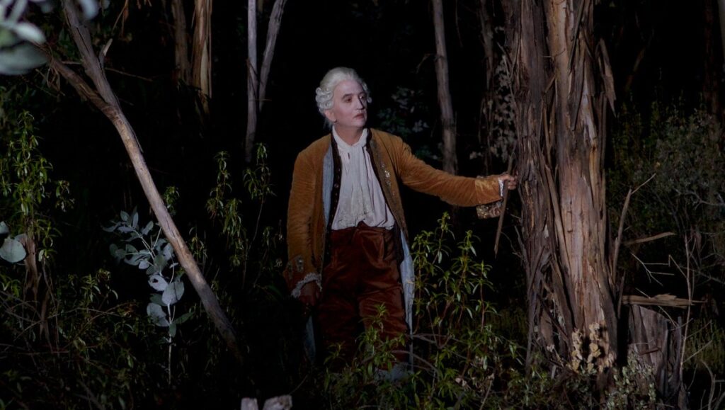 Albert Serra's Liberté: Aristocrat in 1700s attire stands in a dark forest. Film still.