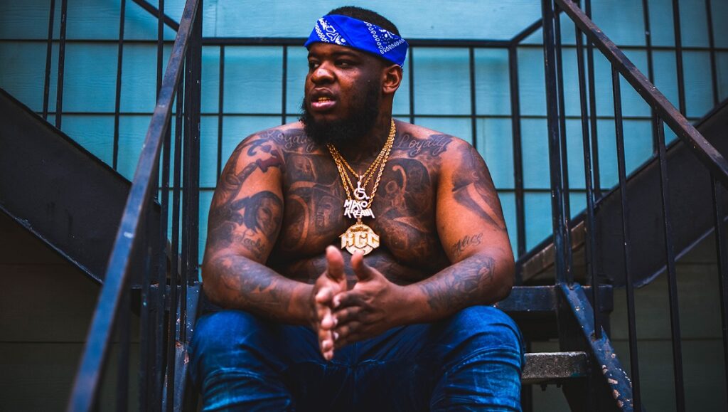 Maxo Kream | Brandon Banks Maxo Kream, the Brandon Banks rapper, sits on stairs with tattoos and gold chains. Blue bandana.