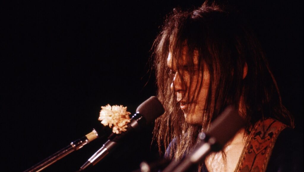 Neil Young performing live. Rock musician with long hair sings into a microphone. Concert photography.