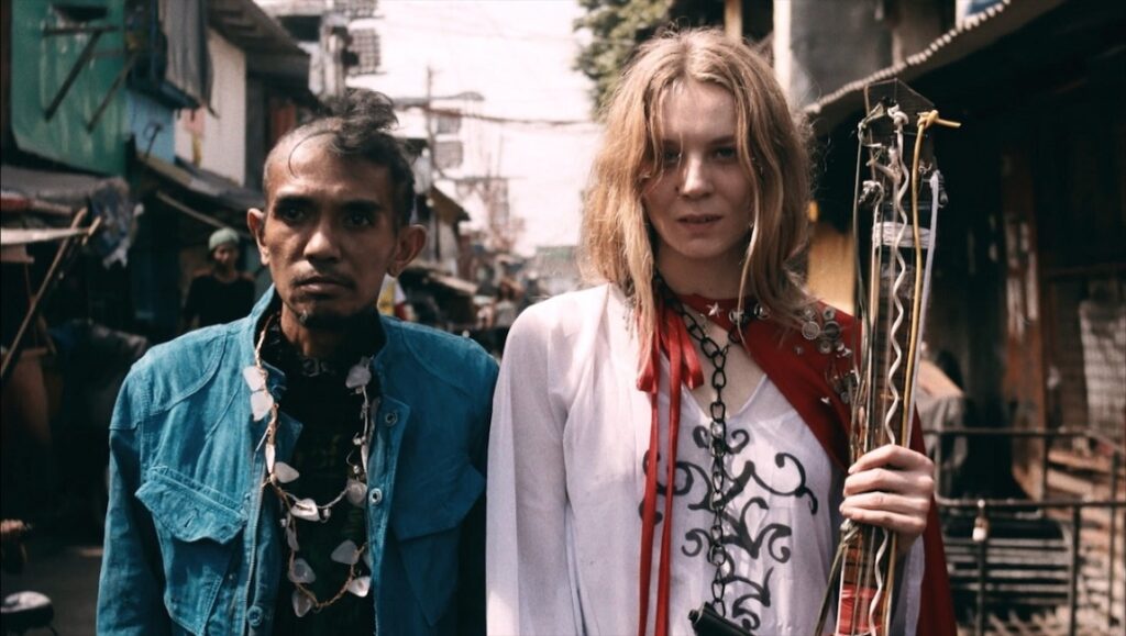 Alexander Kluge & Khavn De La Cruz's Orphea film still. Two people stand in a street. One holds a guitar-like instrument.