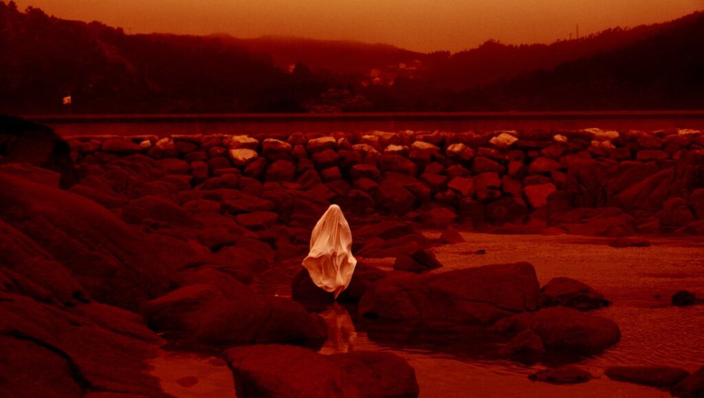 Red Moon Tide by Lois Patiño: Figure draped in fabric on a rocky shore under a red sky. Landscape art.
