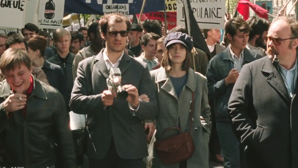 Redoubtable film scene. A crowd of people march with signs. A man holds a camera, and a woman wears a newsboy cap.