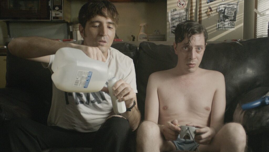 Relaxer movie still. Two men on a couch, one pouring liquid from a jug. Joel Potrykus film.
